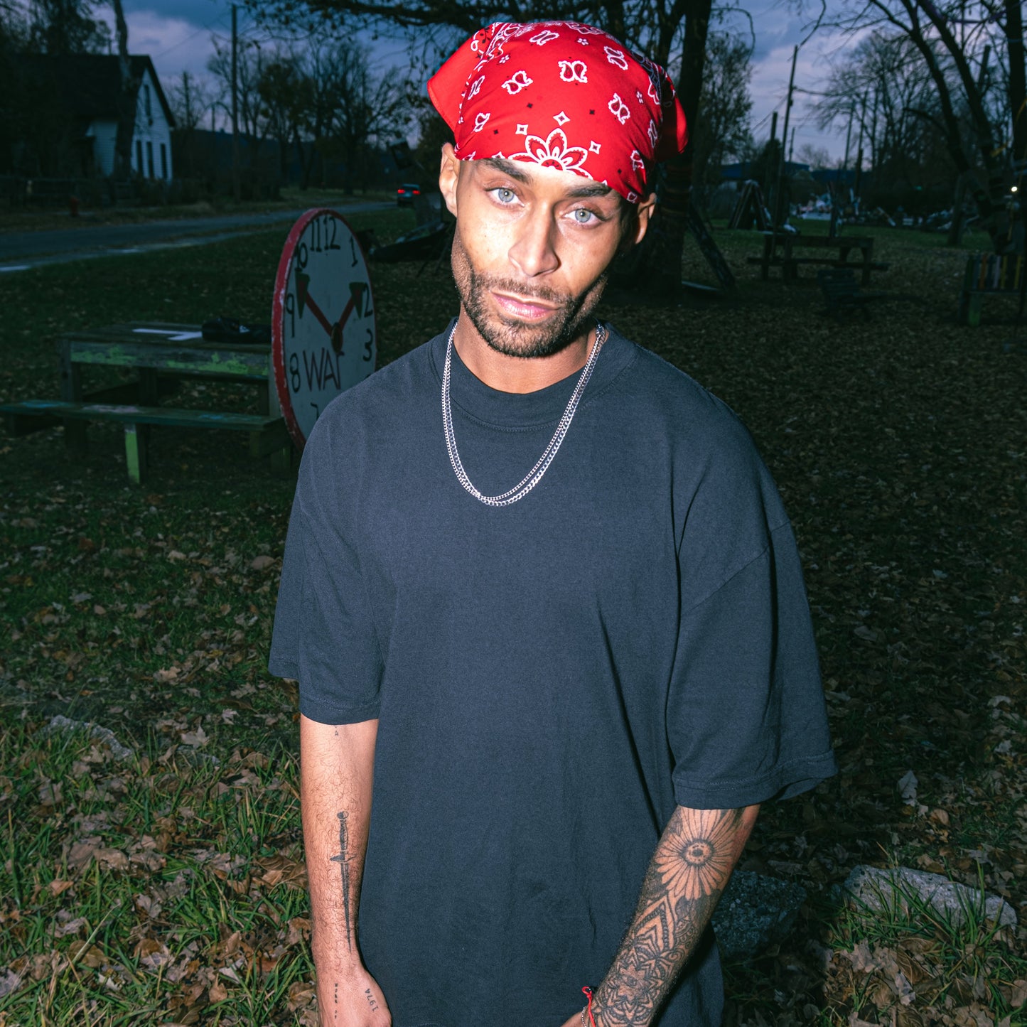 Man wearing a red bandana and black clothing standing outdoors with trees and a road in the background.
