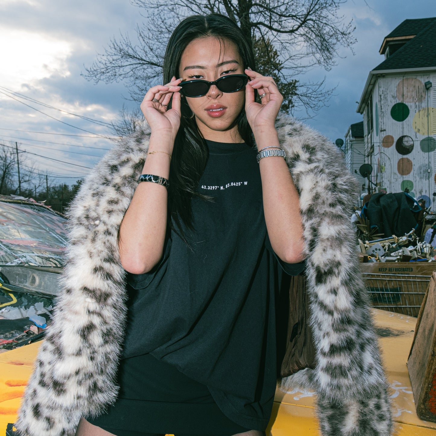 Person wearing a black outfit with a fur coat over shoulders, standing on a yellow taxi with a colorful building in the background.