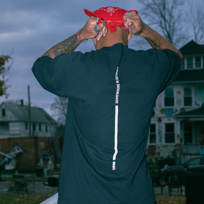 Person wearing a black shirt with text on the back, standing outdoors with a red bandana on their head.