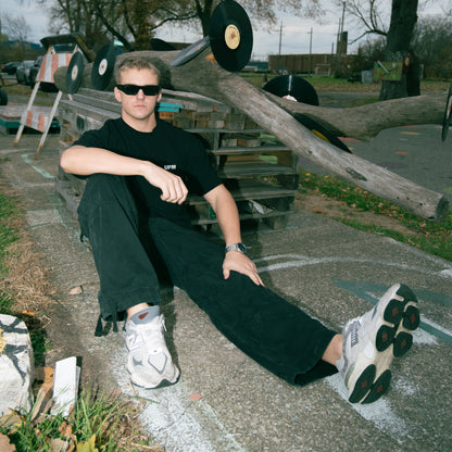 Person sitting on a concrete ledge wearing sunglasses and black clothing, with a fallen tree and picnic table in the background.