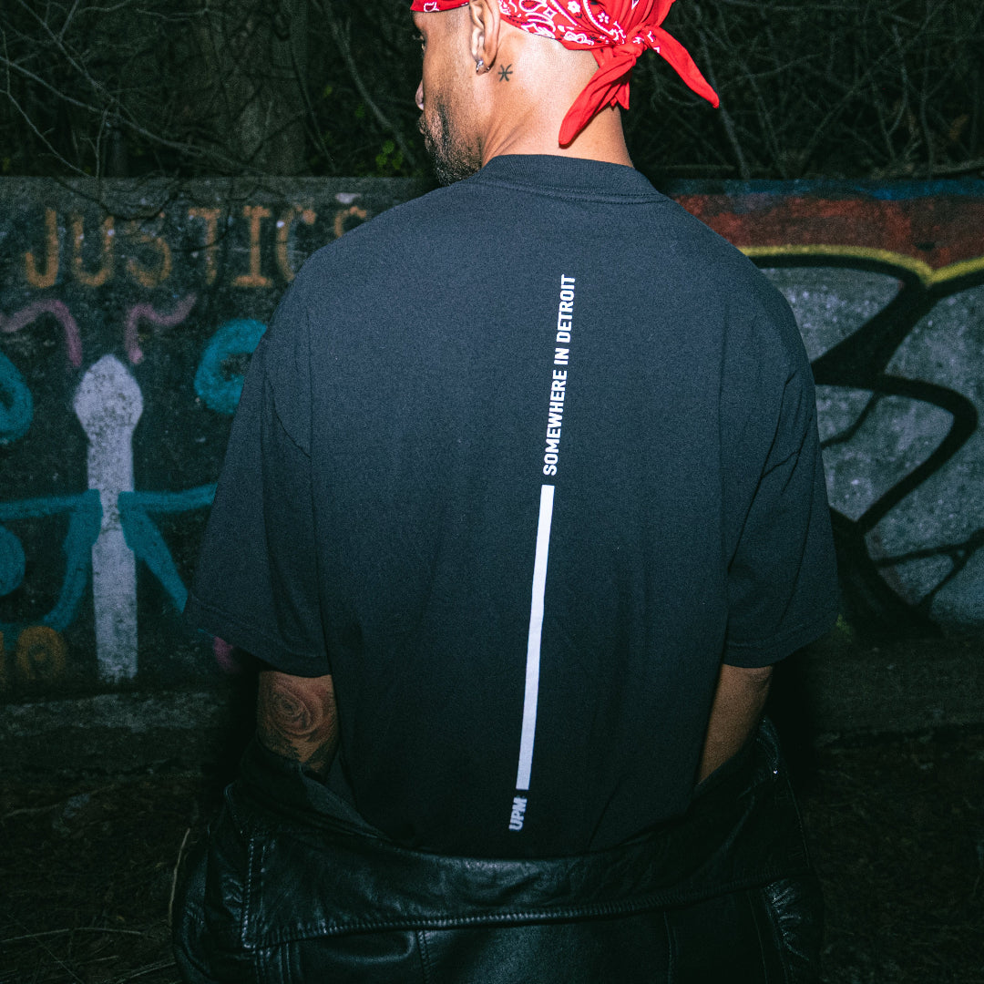 Person wearing a black t-shirt with text on the back, standing in front of a graffiti-covered wall.