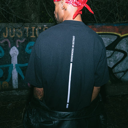 Person wearing a black t-shirt with text on the back, standing in front of a graffiti-covered wall.